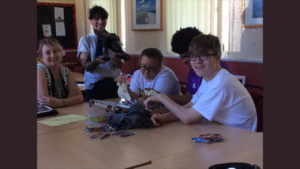Three young people are sat at a table exploring objects while a scriptwriter looks on and a camera person films