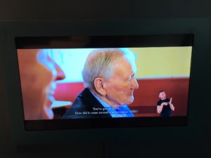 The image shows a tv screen which is showing the film Visions which was created by blind and visually impaired students at St Vincents School for Sensory Impairment. On the screen are two older men who are both laughing plus the subtitles of what is being said and BSL interpretation.