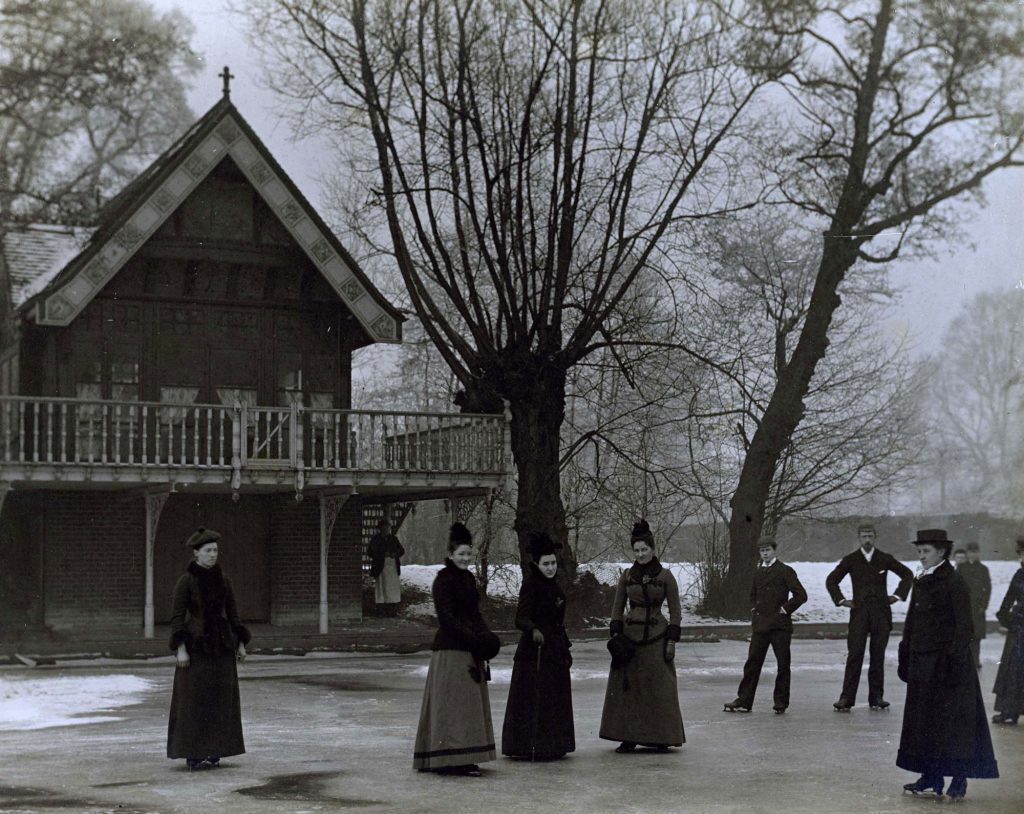 Men and women in Victorian dress on a frozen lake, alongside a building with a wooden balconyLan