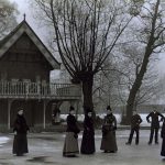 Men and women in Victorian dress on a frozen lake, alongside a building with a wooden balconyLan