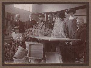Image shows table with half weaved baskets, the completed items on the floor, and a group of weavers standing and sitting around the table.