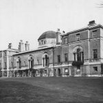 Black and white photo shows imposing long frontage of stately house with arched windows.