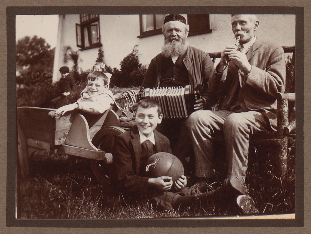 Old men with a whistle and accordion sit with two young boys with a football and wheelbarrow