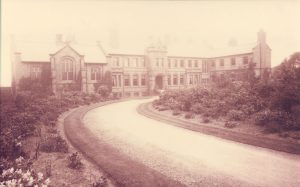 Image shows slightly overexposed black and white photograph of a large Victorian building, with a sweeping drive surrounded by flowerbeds.