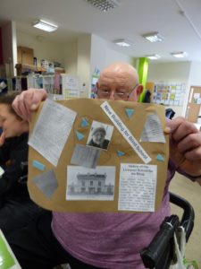 A member of Mencap Liverpool is sat holding up the poster he made which includes information and images from his archive which tell us about his life