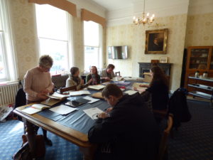 Members of the Liverpool volunteer research group are all sat around a large (seating available for at least twenty people), old fashioned (wood with a leather tabletop) table in the Royal School for the Blind boardroom. They are each exploring text based documents like newspapers, ledgers and letters. On the wall at the head of the table is a framed portrait of Edward Rushon who was one of the origibnal founders of the school.