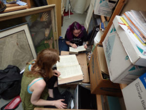 image shows two women sitting on the floor surrounded by files and paintings and looking in reference books