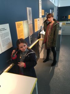 Two young archaeologists stand in The Blind School exhibition holding seakers to their ears to hear the audio description tour