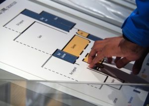 The image shows a hand using fingers to feel a raised architectural floor plan of the school