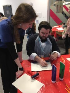 Fae Kilburn is a visually impaired artist and printmaker who is standing next to a table showing Matt Exley, Education Manager at Museum of Liverpool, how to make a print