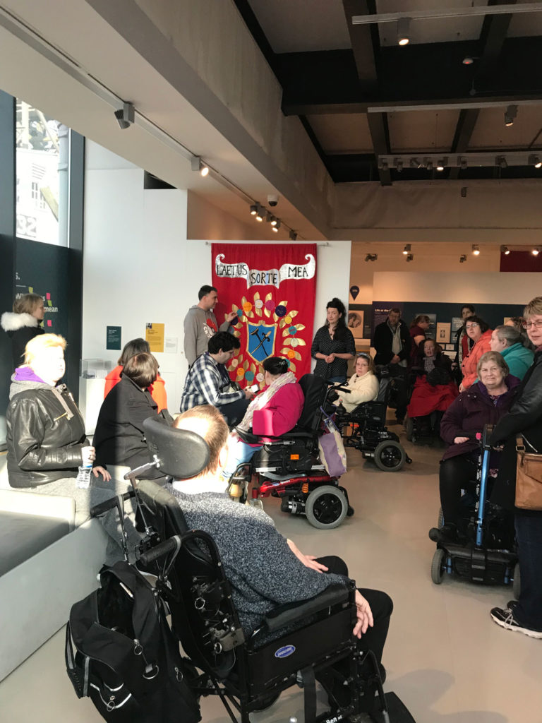 Group of people from Wecil sit in front of the banner made for the Mshed exhibition.