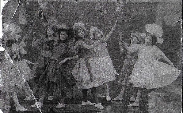 Girls holding ribbons on sticks and dressed in frocks which may be made of paper for dressing up
