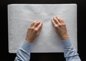 Man's hands on white sheet of paper with raised dots and shapes to indicate building outline and features